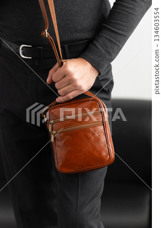 Male model in a charcoal knit crewneck and tailored trousers wearing a cognac leather vertical messenger bag against a white studio background. 134603554