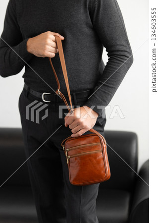 Male model in a charcoal knit crewneck and tailored trousers wearing a cognac leather vertical messenger bag against a white studio background. 134603555