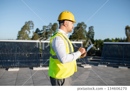 Engineer in protective vest analyzing solar energy installation with gadget Engineer in protective vest analyzing solar energy installation with gadget 134606270