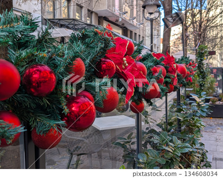 Close up of Christmas garland with red ornaments decorating an outdoor cafe fence in winter highlighting festive city details. Holiday atmosphere, urban decoration, seasonal design and cozy street Close up of Christmas garland with red ornaments decorating an outdoor cafe fence in winter highlighting festive city details. Holiday atmosphere, urban decoration, seasonal design and cozy street 134608034