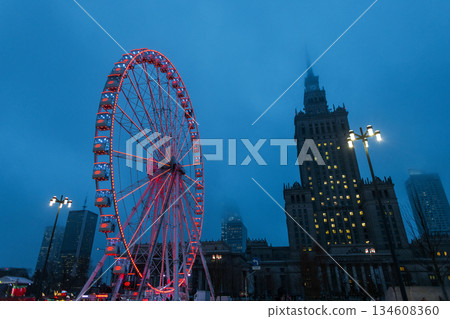 Dramatic low-angle view of an illuminated Ferris wheel in Warsaw at twilight. The glowing red attraction stands against a misty blue sky with the iconic Palace of Culture and Science in the background 134608360