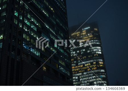 illuminated skyscrapers at night with glowing office windows. glass towers fade into light fog, urban scene that represents corporate life, architecture, finance, and city nightlife. 134608369