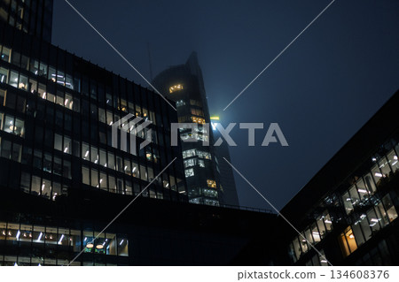 Modern office buildings with glowing windows in a foggy night city. cinematic night shot of sleek glass office towers in a metropolitan business district. Warm lights from the windows contrast Modern office buildings with glowing windows in a foggy night city. cinematic night shot of sleek glass office towers in a metropolitan business district. Warm lights from the windows contrast 134608376