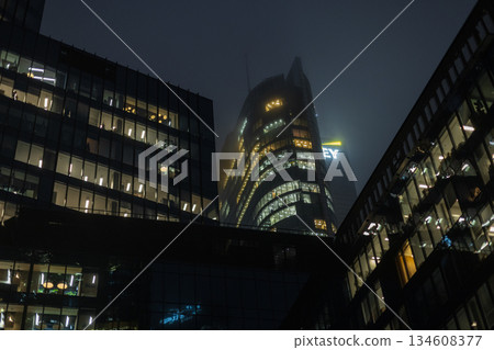 Modern corporate skyscraper with glowing windows in the misty night city. sleek glass skyscraper at night. Warm office lights contrast on a foggy sky, cinematic urban atmosphere. Modern corporate skyscraper with glowing windows in the misty night city. sleek glass skyscraper at night. Warm office lights contrast on a foggy sky, cinematic urban atmosphere. 134608377