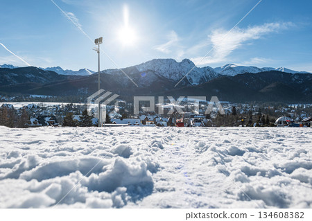 Low-angle view across rough snow toward a winter alpine village nestled below the magnificent, sunlit, snow-covered mountains. Ideal for skiing and travel. Poland, Zakopane, Tatra 134608382