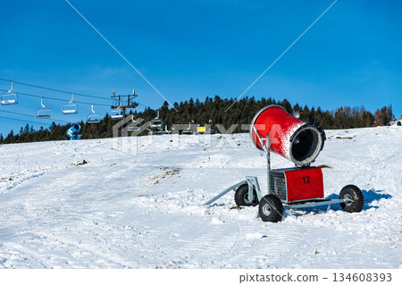 red snow cannon stands ready on a sunny ski slope in Zakopane, Poland, guaranteeing optimal conditions for winter sports. Empty chairlifts ascend the pine-covered mountain against a clear blue sky 134608393