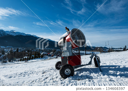 Red snow cannon on a slope preparing the ski run, with a clear view of the Zakopane resort town and the majestic Tatra Mountains against a blue sky.. Winter holidays, extreme mountain recreation 134608397