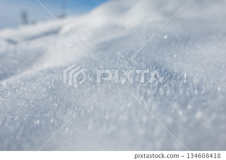 Detailed macro view of untouched snow glistening under bright daylight. The frosty surface reveals natural patterns and subtle contours shaped by wind and cold Detailed macro view of untouched snow glistening under bright daylight. The frosty surface reveals natural patterns and subtle contours shaped by wind and cold 134608418