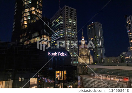 Modern skyscrapers and the iconic Palace of Culture and Science illuminated after dark. prominent Bank Polski sign is visible in the foreground. Urban cityscape, financial district, architect at night Modern skyscrapers and the iconic Palace of Culture and Science illuminated after dark. prominent Bank Polski sign is visible in the foreground. Urban cityscape, financial district, architect at night 134608448