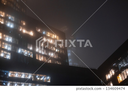 massive high-rise illuminated by thousands of watts, showcasing the immense energy consumption of a modern megacity. Foggy night cityscape with illuminated office building windows glowing 134609474