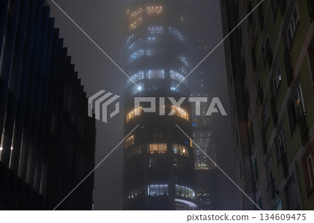 glass facade of a corporate skyscraper glowing with electrical light under heavy weather conditions. Foggy night cityscape with illuminated office building windows glowing through dense mist. glass facade of a corporate skyscraper glowing with electrical light under heavy weather conditions. Foggy night cityscape with illuminated office building windows glowing through dense mist. 134609475