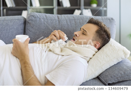 Unwell man with virus lying under blanket on sofa, drinking tea, using tissues Unwell man with virus lying under blanket on sofa, drinking tea, using tissues 134609479