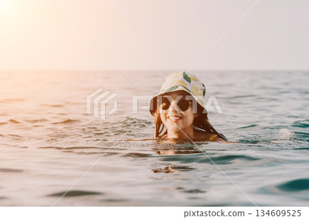 Woman Sunglasses Ocean - Smiling woman wearing sunglasses and a bucket hat swims in the ocean on a sunny day. 134609525