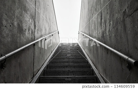Industrial Staircase Metal. A stark, angular shot looking up a utilitarian metal staircase with perforated treads. The stairs ascend alongside 134609559