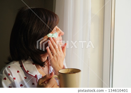 Woman applying eye patches and drinking tea during morning self-care routine by window 134610401
