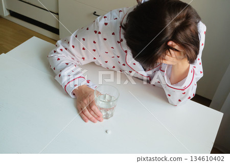 Woman wearing pajamas experiencing headache, sitting at table with medicine and water 134610402