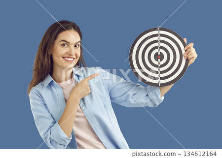 Happy positive young woman sets work goal and points at bullseye on shooting target 134612164