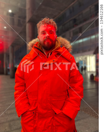 Man in red winter parka standing in urban transit space at night. Travel pause, calm presence, modern city movement and personal journey within architectural environment. 134612166