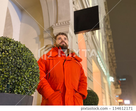 Man in red winter parka standing near building facade at night. Urban exploration, upward gaze, personal journey and quiet anticipation within modern city environment. 134612171