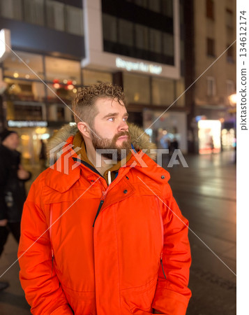 Portrait of man in red parka on city street at night. Urban lifestyle, winter fashion, thoughtful mood, individuality and calm presence within illuminated evening environment. 134612174
