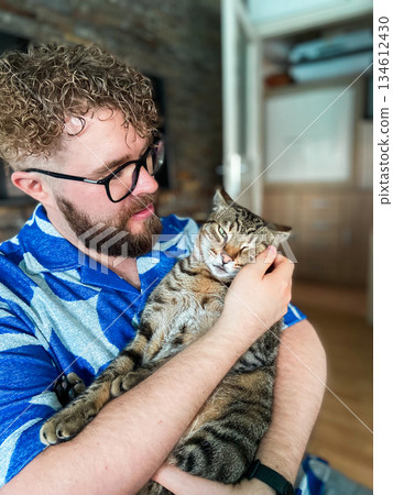 Man holding tabby cat in arms inside home interior. Emotional connection, pet companionship and calm domestic lifestyle expressing care, trust and everyday affection. 134612430