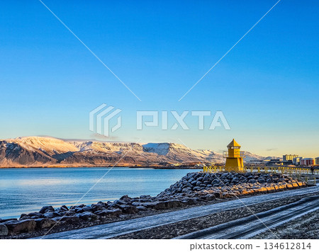 Yellow Lighthouse on Rocky Coast with Snowy Mountains and Blue Sky Yellow Lighthouse on Rocky Coast with Snowy Mountains and Blue Sky 134612814