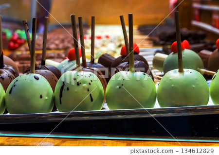 A row of green chocolate coated apples on sticks displayed at a christmas market sweets stall, with colorful treats visible in the background. A row of green chocolate coated apples on sticks displayed at a christmas market sweets stall, with colorful treats visible in the background. 134612829