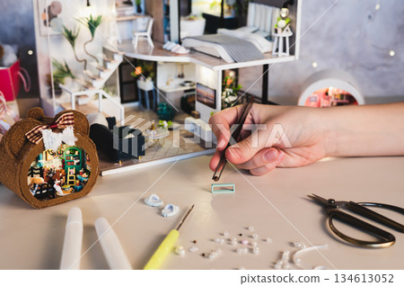 A girl uses tweezers to assemble decorations for a miniature dollhouse. 134613052