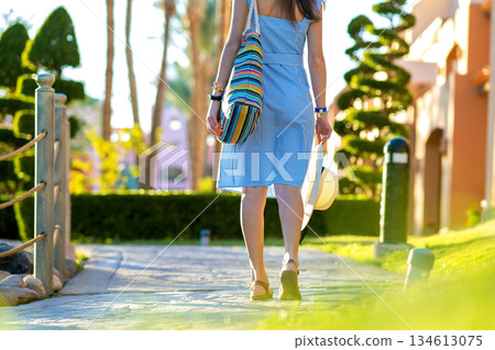 Young woman wearing light blue summer dress holding fashionable shoulder bag and yellow straw hat standing outside enjoying warm weather in summer park. Young woman wearing light blue summer dress holding fashionable shoulder bag and yellow straw hat standing outside enjoying warm weather in summer park. 134613075