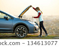 Young woman opening bonnet of broken down car having trouble with her vehicle. Female driver standing near auto with popped up hood. 134613077