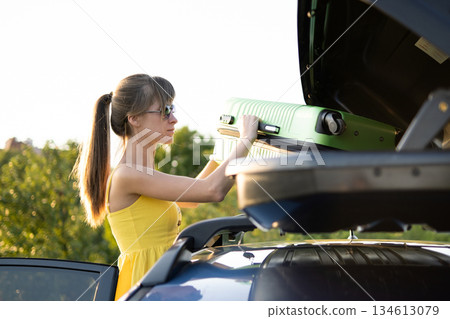 Young woman driver taking green suitcase from car roof rack. Travel and vacations concept. 134613079