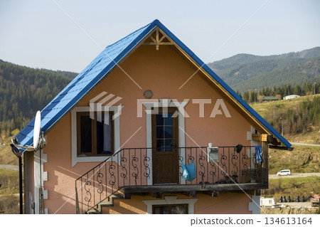 Two-storied residential cottage house top with shingle blue roof, plastered pink walls, forged balcony and outside staircase in ecological mountain area on blue sky copy space background. Two-storied residential cottage house top with shingle blue roof, plastered pink walls, forged balcony and outside staircase in ecological mountain area on blue sky copy space background. 134613164