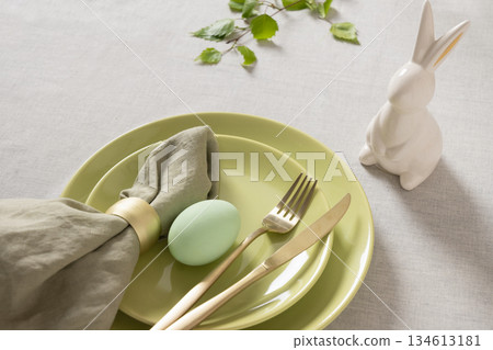 Minimal Easter place setting with pastel egg, green ceramic plates, golden cutlery and white bunny figurine on neutral tablecloth, spring holiday concept. Minimal Easter place setting with pastel egg, green ceramic plates, golden cutlery and white bunny figurine on neutral tablecloth, spring holiday concept. 134613181