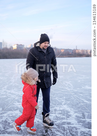 A father and his little daughter in a red winter jumpsuit skate in the fresh frosty air on the frozen surface of the lake 134613189