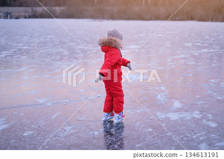 A girl in a bright red winter jumpsuit skates on the ice rink of a frozen lake at sunset 134613191