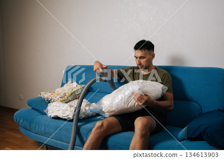 Focused young man sitting on sofa using vacuum cleaner to suck air out of storage bag containing bedding, reducing storage space and protecting from dust and moisture. Concept of household chores. Focused young man sitting on sofa using vacuum cleaner to suck air out of storage bag containing bedding, reducing storage space and protecting from dust and moisture. Concept of household chores. 134613903