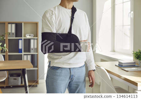 Close-up photo of a man's arm in a cast standing at home or at office wearing sling. Close-up photo of a man's arm in a cast standing at home or at office wearing sling. 134614113