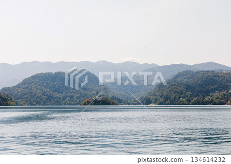 Idyllic view of Lake Bled surrounded by green hills and distant peaks, reflecting in the peaceful water on a clear day. Old church on the island Idyllic view of Lake Bled surrounded by green hills and distant peaks, reflecting in the peaceful water on a clear day. Old church on the island 134614232