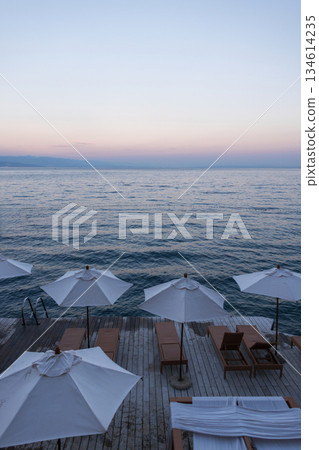 Relaxing summer holiday scene with beach chairs and parasols on a wooden deck by the calm sea. Seaside view in Croatia with white parasols and wooden sunbeds at sunset, overlooking the calm Adriatic 134614235
