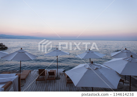Seaside view in Croatia with white parasols and wooden sunbeds at sunset, overlooking the calm Adriatic Sea and distant mountains. Perfect summer vacation and travel background 134614238