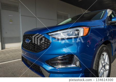 Blue crossover vehicle parked outdoors near parking zone, modern headlight and grille detail. Compact blue crossover SUV car standing in urban parking space outdoors. Blue crossover vehicle parked outdoors near parking zone, modern headlight and grille detail. Compact blue crossover SUV car standing in urban parking space outdoors. 134614300