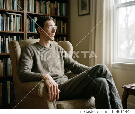 individual with Marfan syndrome is seated in a chair next to a window. The room contains bookshelves filled with various books and there is natural light coming in. 134615443