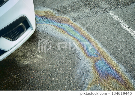 Car leaking engine oil, creating a colorful iridescent rainbow pattern on dark asphalt. 134619440
