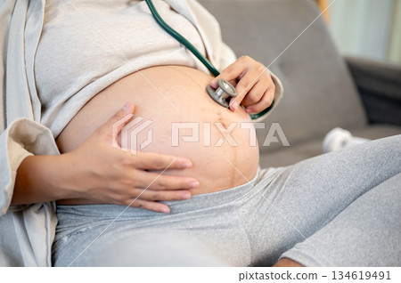 Close up of pregnant woman using stethoscope on belly listening to fetal heartbeats sitting on sofa Close up of pregnant woman using stethoscope on belly listening to fetal heartbeats sitting on sofa 134619491