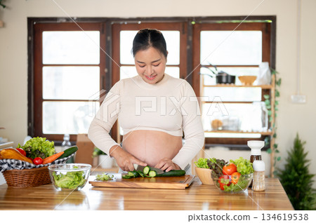 Pregnant asian woman cutting a cucumber with knife on chopping board preparing her food in kitchen. Pregnant asian woman cutting a cucumber with knife on chopping board preparing her food in kitchen. 134619538