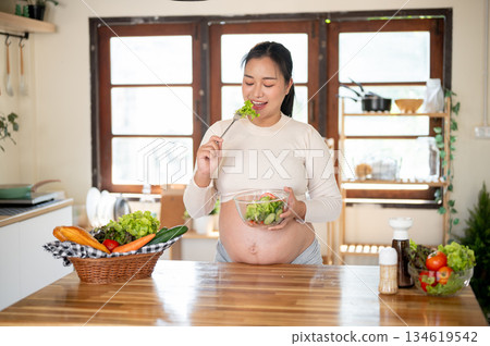 Pregnant asian woman holding glass bowl enjoy eating a vegetables salad she made at kitchen counter. Pregnant asian woman holding glass bowl enjoy eating a vegetables salad she made at kitchen counter. 134619542