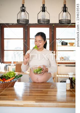 Pregnant asian woman holding fork enjoy eating vegetables salad she made at kitchen cooking counter. Pregnant asian woman holding fork enjoy eating vegetables salad she made at kitchen cooking counter. 134619543