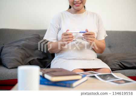 Pregnant asian woman is smiling and looking at pregnancy test kit in hand sitting on sofa in house. Pregnant asian woman is smiling and looking at pregnancy test kit in hand sitting on sofa in house. 134619558