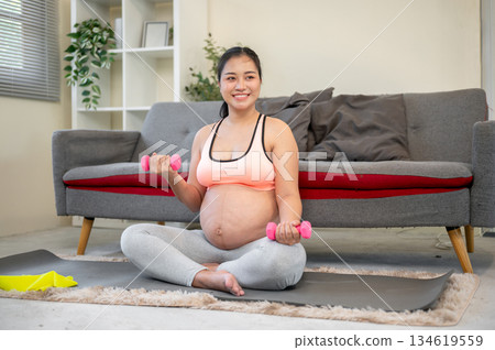 Pregnant asian woman with her bare belly lifting dumbbell while sitting on rubber mat in the house's living room. 134619559