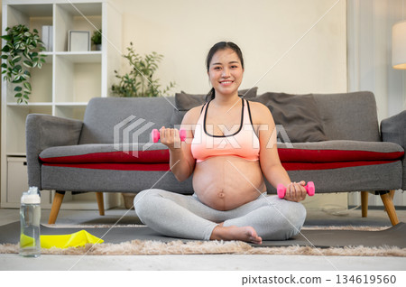 Pregnant asian woman with bare belly smiling as lifting dumbbell while sitting on mat in living room 134619560